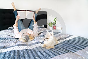 Blonde hair woman sitting on cozy bed with dog, holding open book and reading. Lying woman relaxing on sofa at home