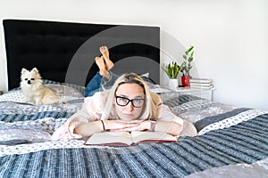 Blonde hair woman sitting on cozy bed with dog, holding open book and reading. Lying woman relaxing on sofa at home