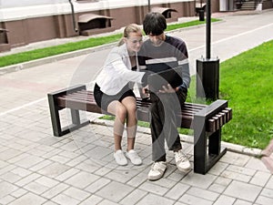 Blonde-haired adult girl in glasses, smiling, shows something on the laptop to a dark-haired man, sitting on a bench. People