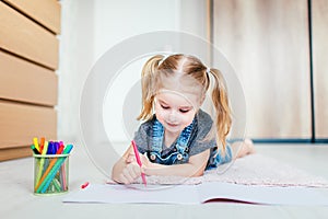 Blonde happy little girl with two ponytales drawing and writing lying on the floor at home. Preschool education, early learning