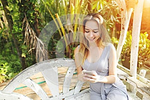 Blonde happy woman uses smartphone for work on sunny day, background of sunshine green palms in Thailand, Phuket travel