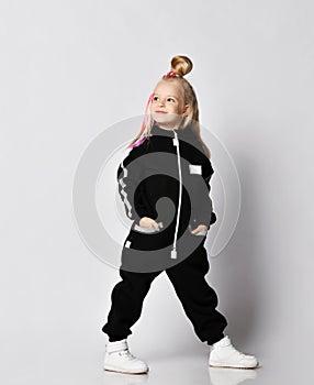 Blonde kid baby girl with braids wearing stylish black jumpsuit and sneakers stands side to camera and looks back and up