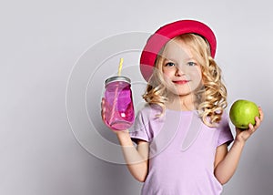 Blonde kid in red hat and purple blouse. She smiling, holding pink cocktail bottle and green apple, posing isolated on white