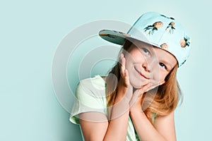 Blonde kid in t-shirt and cap with pineapples print. She smiling and touching her face, posing on turquoise studio background.