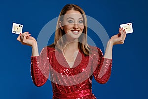 Blonde lady with make-up, in red sequin dress is smiling, showing two aces, posing on blue background. Gambling, poker