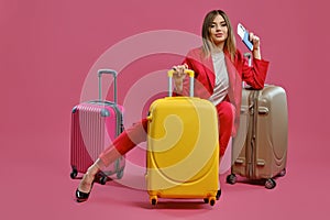 Blonde lady in red pantsuit, white blouse, black heels. She smiling, sitting among colorful suitcases, holding passport