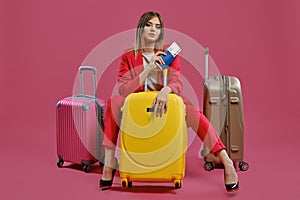 Blonde lady in red pantsuit, white blouse, high black heels. She sitting among three colorful suitcases, holding