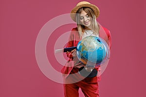 Blonde lady in straw hat, white blouse and red pantsuit. She is smiling, holding globe, passport and ticket, posing