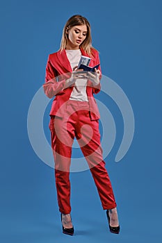 Blonde lady in white blouse, red pantsuit, high black heels. She is looking at her passport and ticket while posing on