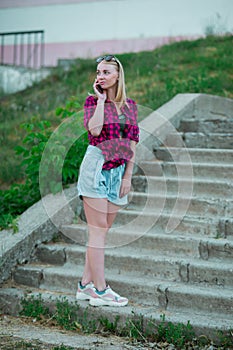 Blonde in light blue high-rise shorts, red plaid shirt, sneakers standing on the stairs outside, looks to the left, talking on a