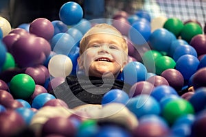 Blonde little boy lying on multi coloured plastic balls in big dry paddling pool in playing centre. Smiling at camera