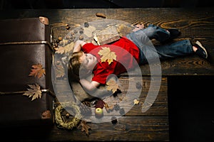 Blonde little boy resting with leaf on stomach lies on wooden floor in autumn leaves. Cute little child boy are getting