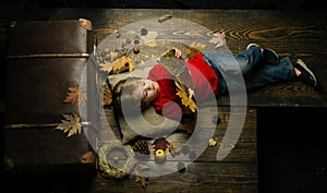 Blonde little boy resting with leaf on stomach lies on wooden floor in autumn leaves. Little cute boy lies on a wooden