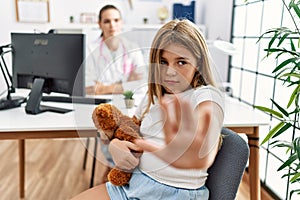 Blonde little girl at pediatrician clinic with female doctor with open hand doing stop sign with serious and confident expression,