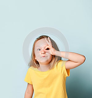 Blonde little girl in yellow t-shirt. Showing ok sign and looking through it while posing against turquoise studio background.
