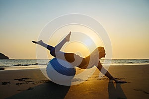 Blonde with long hair makes Pilates on the beach during sunset against the sea. Young flexible happy woman doing fitness exercises