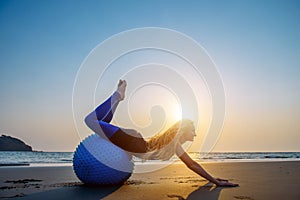 Blonde with long hair makes Pilates on the beach during sunset against the sea. Young flexible happy woman doing fitness exercises