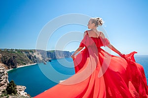 Blonde with long hair on a sunny seashore in a red flowing dress, back view, silk fabric waving in the wind. Against the
