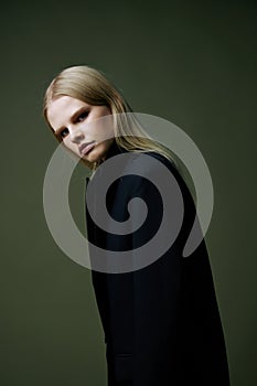 Blonde looks over her shoulder in a black jacket posing half-turned in the studio on a green background. The concept of