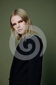 Blonde looks over her shoulder in a black jacket posing half-turned in the studio on a green background. The concept of