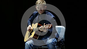 Blonde man playing acoustic guitar on stage at spotlight, unplugged performance.