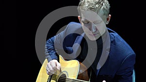 Blonde man playing acoustic guitar on stage at spotlight, unplugged performance.