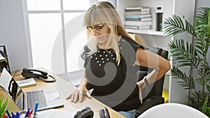 Blonde middle age businesswoman suffering from backache while working in office, touching her aching back, worried expression on