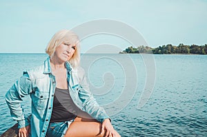 Blonde middle age woman in jeans shirt, sitting on a beach with blue sky background, copy space. Portrait of attractive