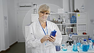 Blonde middle age woman scientist, all smiles as she efficiently uses her smartphone amidst work in the lab