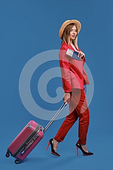Blonde model in hat, white blouse, red pantsuit, high heels. Carrying pink suitcase, showing passport and ticket, posing