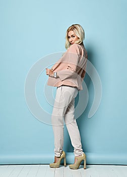 Model with long hair, in beige jacket, white overall, high heels. Smiling, standing against blue studio background. Full length