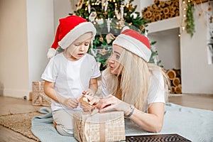 Blonde mom and daughter in Santa hats lie on Mat with laptop and unwrap a Christmas gift. Christmas tree in the back