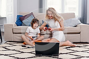 Blonde mother and her little funny son playing video games on console. Happy family time together.