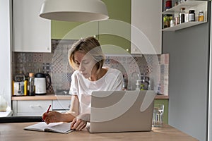 Blonde pensive middle-aged woman working from home using laptop and notebook