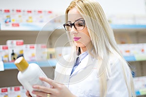 Blonde pharmacist looking at pills and creams in store. Female medical assistant reading on labels