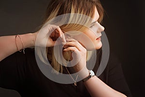 Blonde Plus Size Model on a black background in dark clothes, fastens the earring with two hands