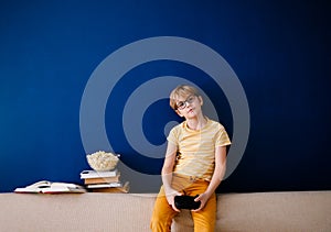 Blonde schoolboy boy plays video games, holds a gamepad, eats popcorn instead of learning lessons on blue background