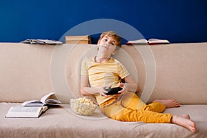 Blonde schoolboy boy plays video games, holds a gamepad, eats popcorn instead of learning lessons on blue background