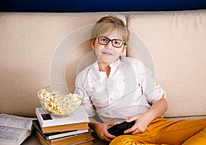 Blonde schoolboy boy plays video games, holds a gamepad, eats popcorn instead of learning lessons on blue background