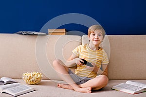 Blonde schoolboy boy plays video games, holds a gamepad, eats popcorn instead of learning lessons on blue background