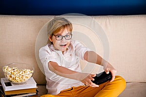 Blonde schoolboy boy plays video games, holds a gamepad, eats popcorn instead of learning lessons on blue background