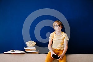 Blonde schoolboy boy plays video games, holds a gamepad, eats popcorn instead of learning lessons on blue background