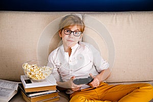 Blonde schoolboy boy plays video games, holds a gamepad, eats popcorn instead of learning lessons on blue background