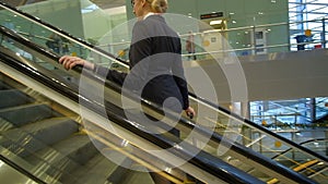 Blonde stewardess rides up on escalator with suitcase at airport