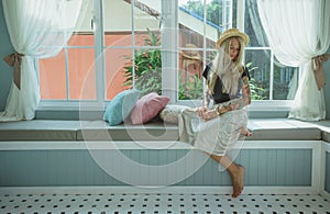 Blonde in a straw hat sits on the couch by the window. the image of a modern girl. Model with tattoos