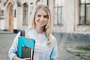 Student girl is smiling and holding a folder and a notebook in her hands on a university background. Girl is taking exams