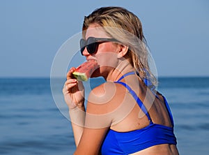 A blonde in sunglasses eats a watermelon by the sea. A juicy watermelon in the hands of a woman