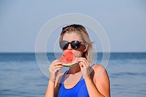 A blonde in sunglasses eats a watermelon by the sea. A juicy watermelon in the hands of a woman