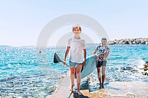 Blonde teenage son with father surfers smiling and carrying the stand-up paddleboard with a paddle on the bright sunny day noon.