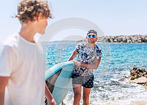 Blonde teenage son gazing to smiling father surfer carrying the surfing board on the bright sunny day noon. Active family summer